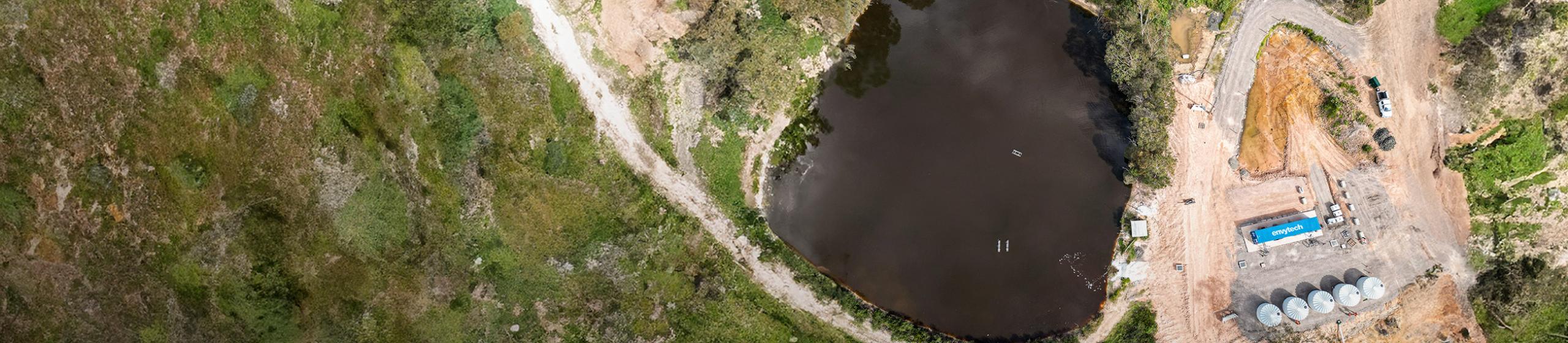 Flygbild över en anläggning för vattenrening vid en sjö, omgiven av grönska och grusvägar. Flera tankar och en blå container syns på anläggningen.